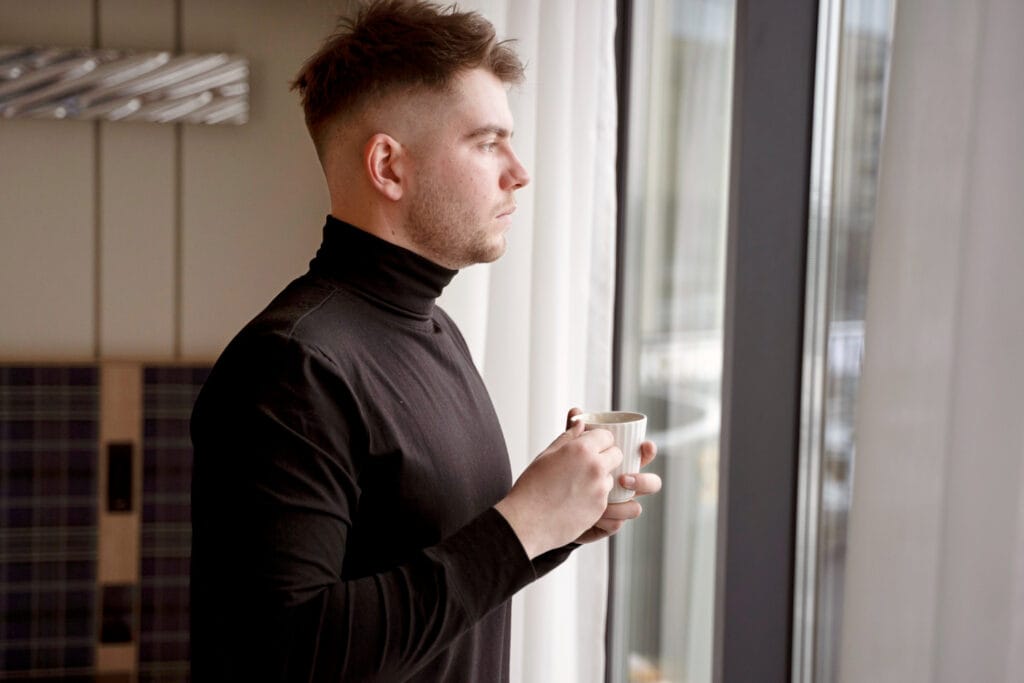 Homem introspectivo olhando pela janela, representando uma origem emocional profunda ligada ao toque evitado.